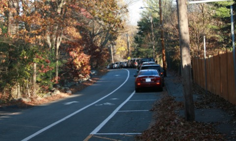 This figure is an image showing a line of private autos waiting on Bow Street to pick up students at dismissal time. A bicycle lane has been striped next to the parked cars.